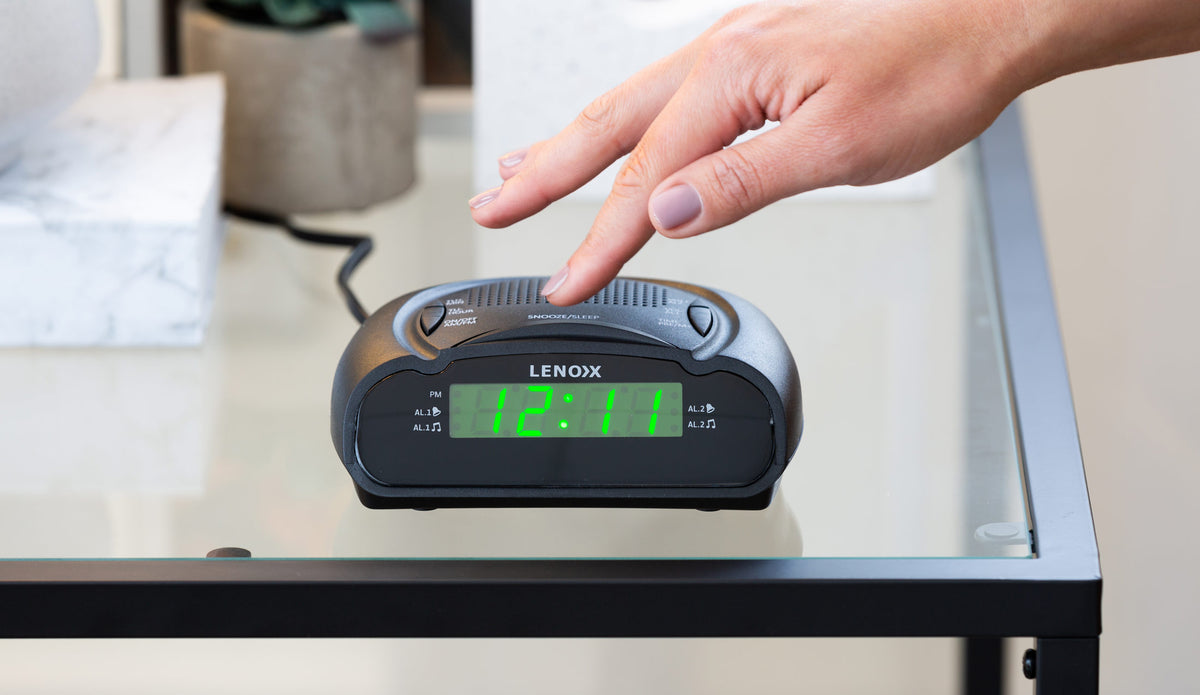 Black rounded AM/FM Alarm Clock & Radio with Large Green LED Time Numbering on a glass table with a hand pressing the snooze button.
