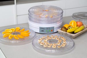 Round food dehydrator in a kitchen with trays of bananas and oranges next to it.