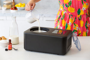 Person pouring liquid into a black ice cream maker on a kitchen counter.