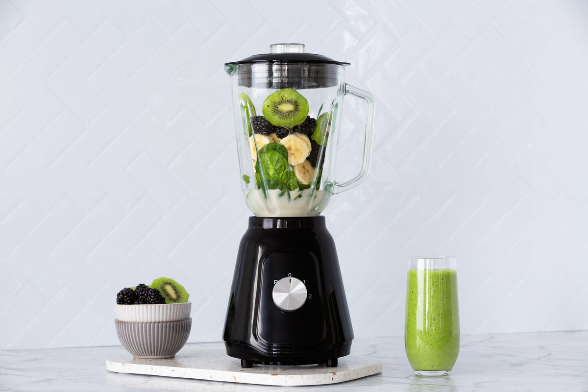 Black table blender with its glass jug filled with kiwi, blackberries and bananas and a green smoothie in a glass next to it.