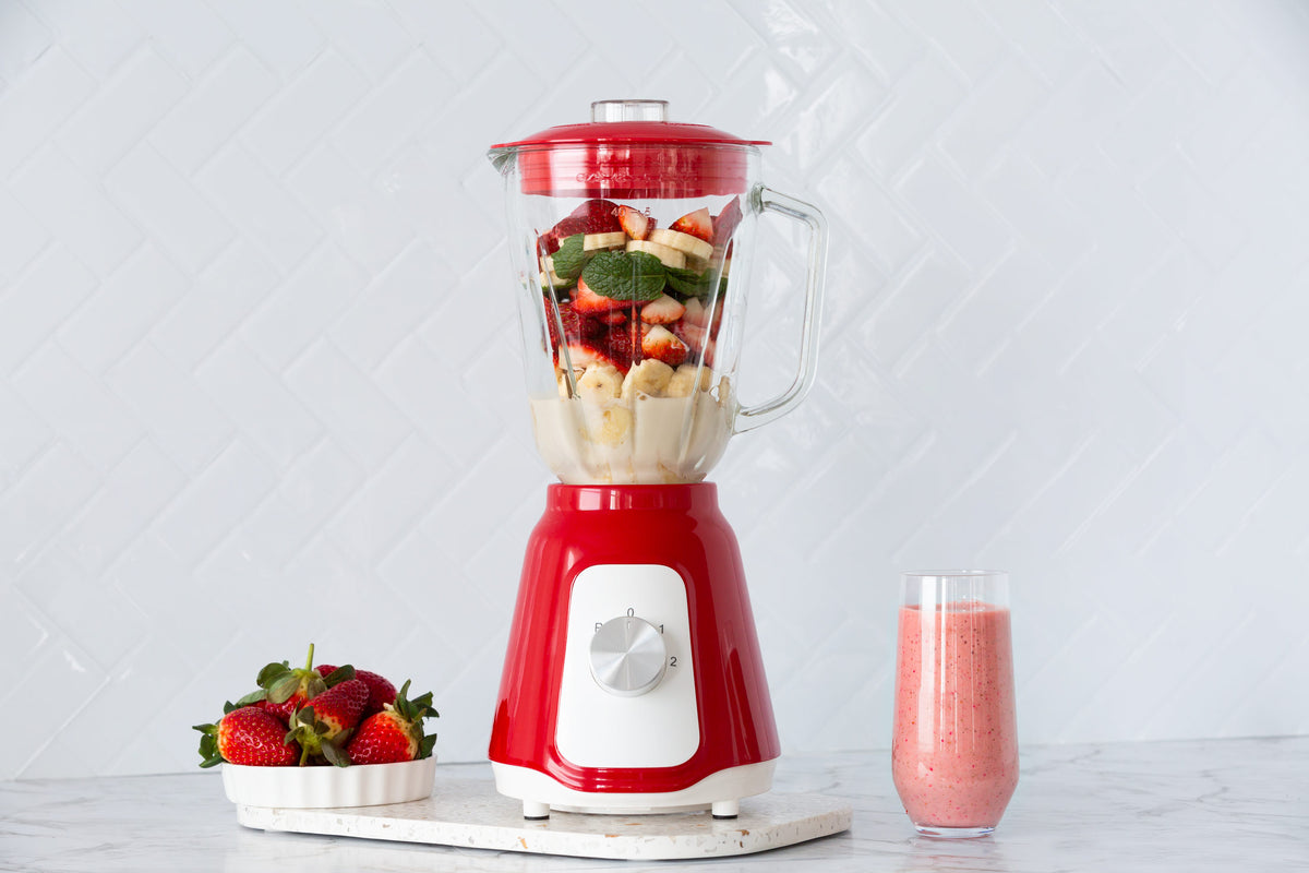 Red table blender with its glass jug filled with strawberries and bananas and a smoothie in a glass next to it.