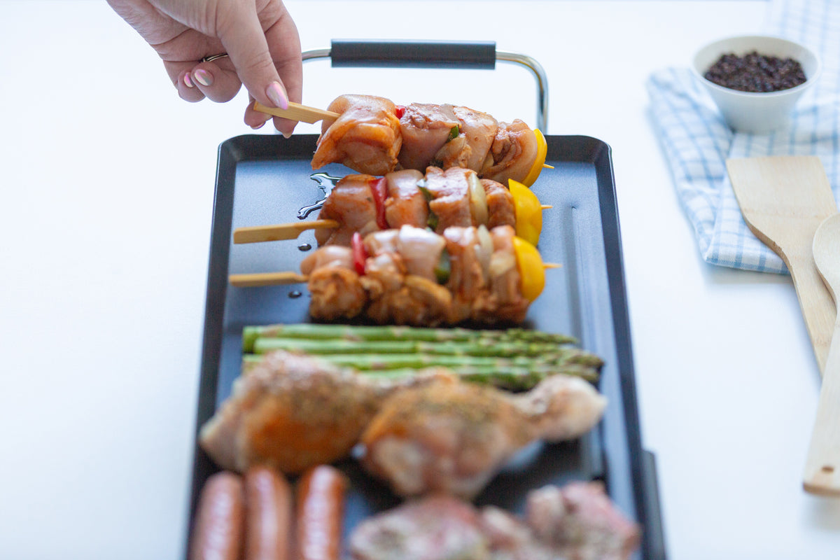 Person preparing skewered food on the electric teppanyaki grill with ingredients like sausages and vegetables.