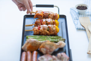Person preparing skewered food on the electric teppanyaki grill with ingredients like sausages and vegetables.