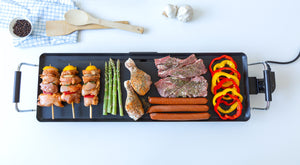 Electric teppanyaki grill with skewers, vegetables, and meats on a white table.

