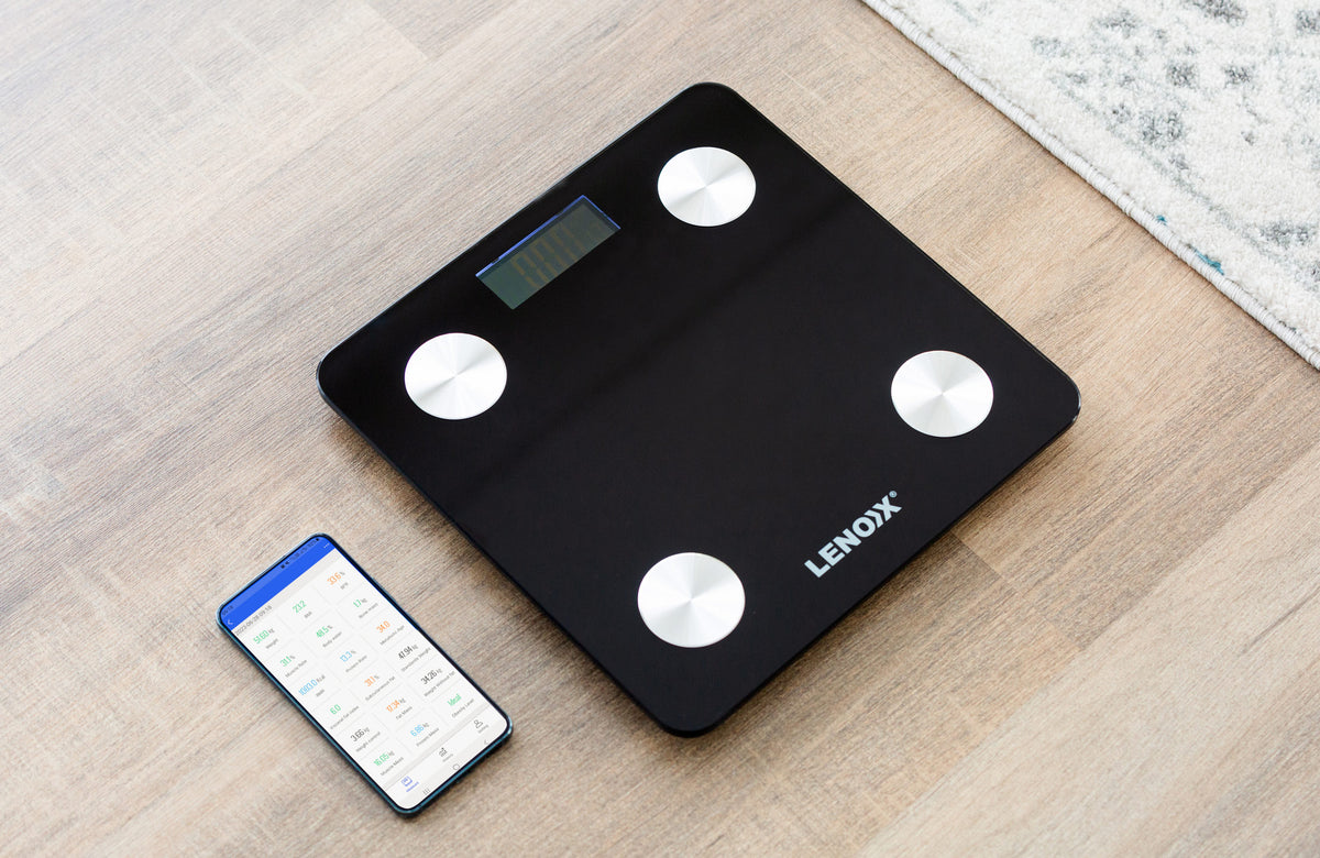 Smart scale on a wooden floor with the app connected displayed on the phone next to the scale.