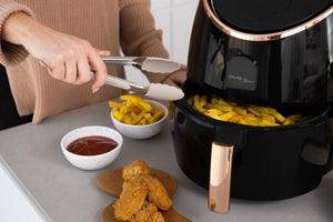 AF720 Air Fryer with the cooking basket open. Woman's hand holding tongs, she's about to pick the hot chips out of the drawer.