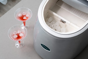 Close up of the Ice Cube Maker with the lid open. 2 cocktails with ice cubes standing next to it.