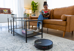 RV2100 robot vacuum on a light carpet in a modern living room. A young woman in the background reads a book while seating on a sofa.