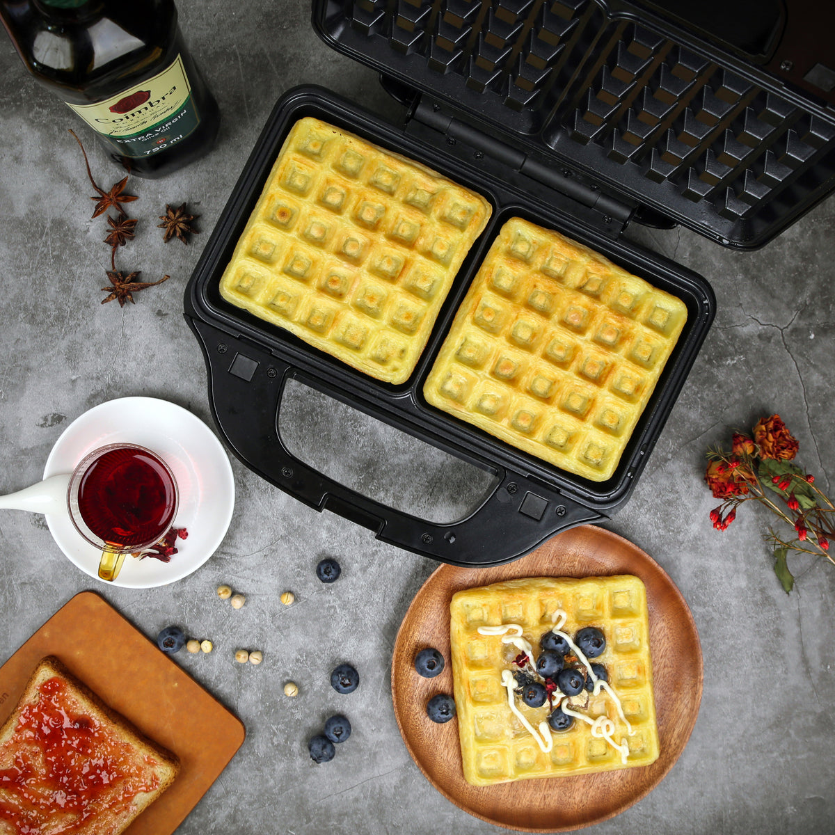 Top view of freshly made waffles in the sandwich press and on plates next to it - served with jam and fresh blueberries.